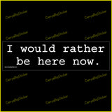 Bumper sticker or magnetic bumper sticker says, I would rather be here now. White lettering on a black background.